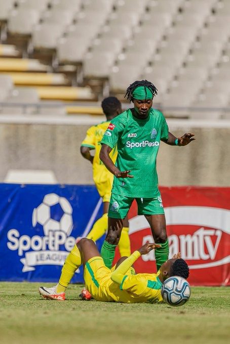 Soccer player in a green uniform stands over a fallen opponent in yellow on a grassy field.