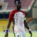 Soccer player in a white and red jersey with a captain's armband walking on a stadium field.
