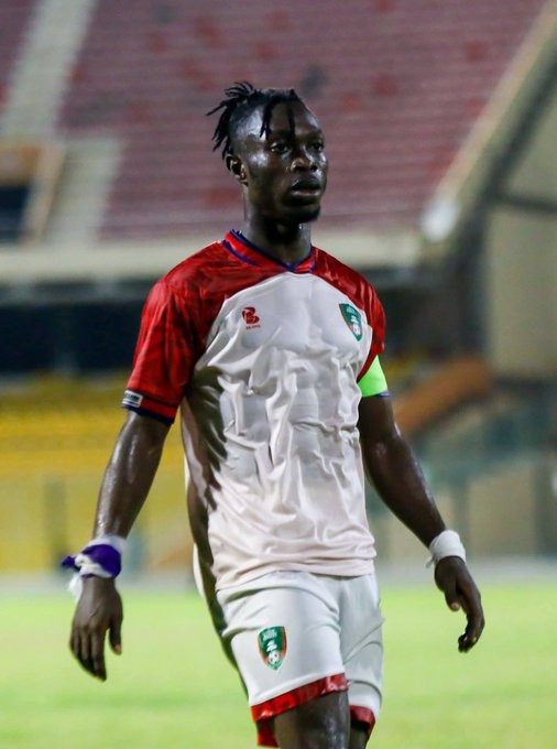 Soccer player in a white and red jersey with a captain's armband walking on a stadium field.
