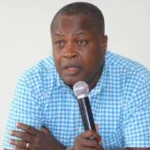 Man in a blue checkered shirt speaks into a handheld microphone at a table, with water bottles in view.
