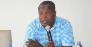Man in a blue checkered shirt speaks into a handheld microphone at a table, with water bottles in view.