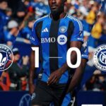 Soccer player in blue and black striped jersey walking on the field with a bold 1–0 score overlay, crowd in the background, team crests and sponsor logos visible.