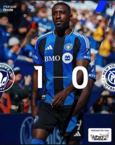 Soccer player in blue and black striped jersey walking on the field with a bold 1–0 score overlay, crowd in the background, team crests and sponsor logos visible.