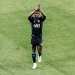 Soccer player in black kit clapping above his head on a green pitch, celebrating a goal on the field.