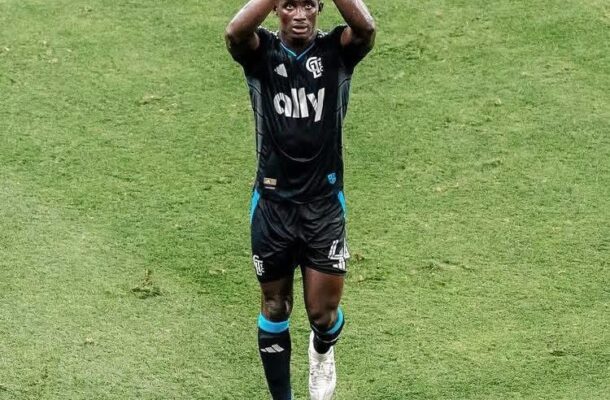 Soccer player in black kit clapping above his head on a green pitch, celebrating a goal on the field.