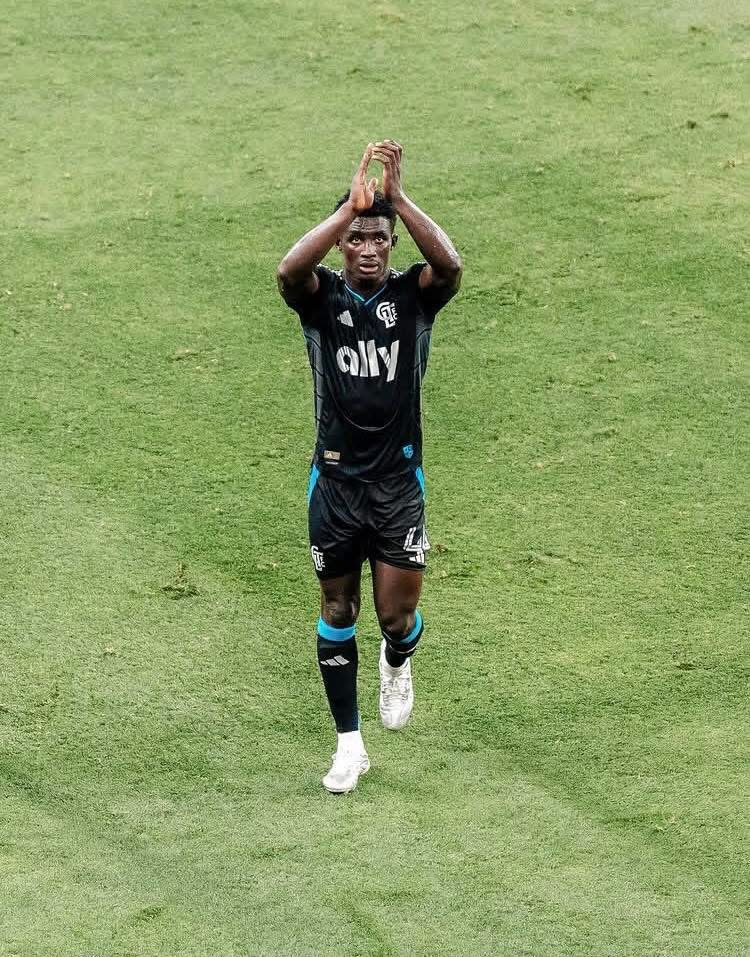 Soccer player in black kit clapping above his head on a green pitch, celebrating a goal on the field.