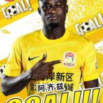 Male soccer player in a bright yellow jersey pointing to the team crest with a 'GOAL!!!' background banner, celebrating a goal.