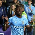 Soccer player in light-blue kit raises both arms in celebration as fans cheer behind him.