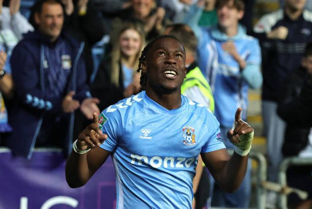 Soccer player in light-blue kit raises both arms in celebration as fans cheer behind him.