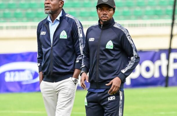 IMG_0299 | Ghanaguardian.com Two men in matching navy tracksuits walk on a football pitch, stadium seats in background; one wears a light gray cap, the other a black cap with AZO logo on jackets.