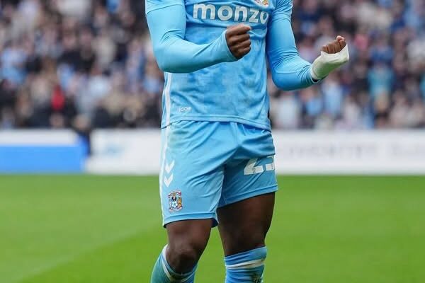 Footballer in a light-blue kit celebrates on a grass pitch with fists clenched and mouth open, crowd blurred in the background.
