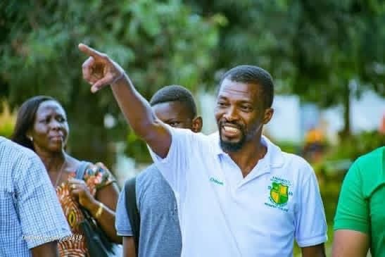 Man in a white polo with a green crest points toward something while others stand behind him in a park-like outdoor setting.