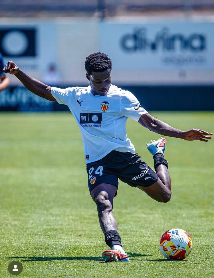 Soccer player in a white jersey and dark shorts kicking a colorful ball on a green grass field during practice.