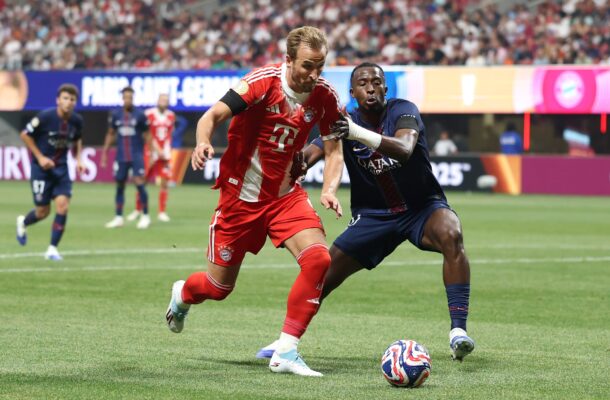 Two soccer players battle for the ball during a match, one in red Bayern kit and the other in dark blue, on a green field with a crowded stands backdrop.