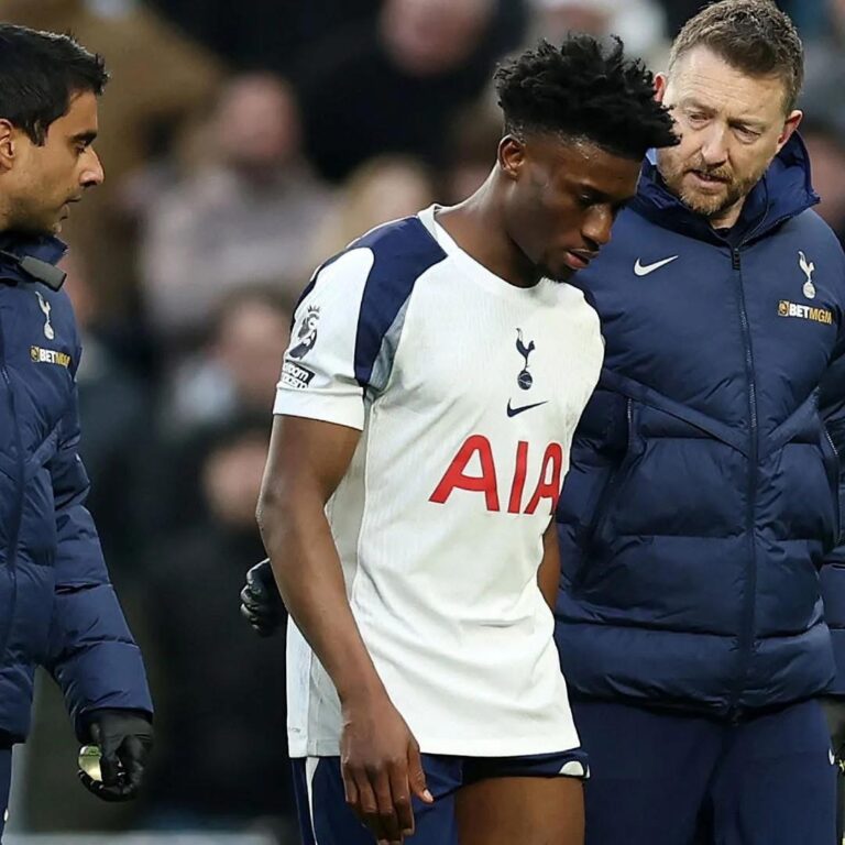 Tottenham player in a white kit is escorted off the pitch by two staff members in navy jackets.