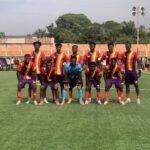 Soccer team lined up for a group photo on a grassy field, players in red-yellow striped jerseys and purple shorts with a blue-clad goalkeeper center.