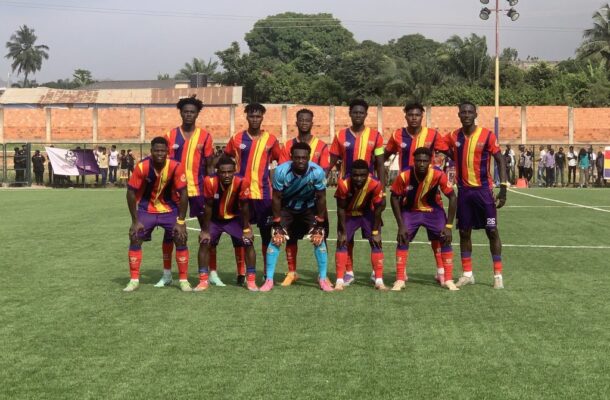 Soccer team lined up for a group photo on a grassy field, players in red-yellow striped jerseys and purple shorts with a blue-clad goalkeeper center.