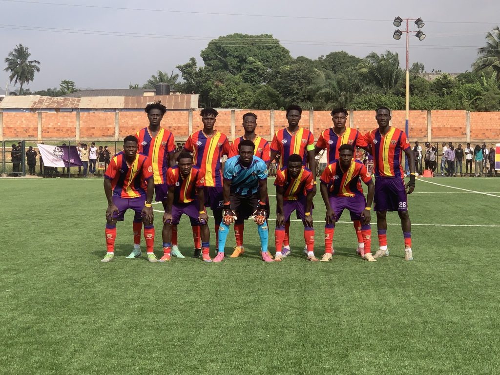 Soccer team lined up for a group photo on a grassy field, players in red-yellow striped jerseys and purple shorts with a blue-clad goalkeeper center.