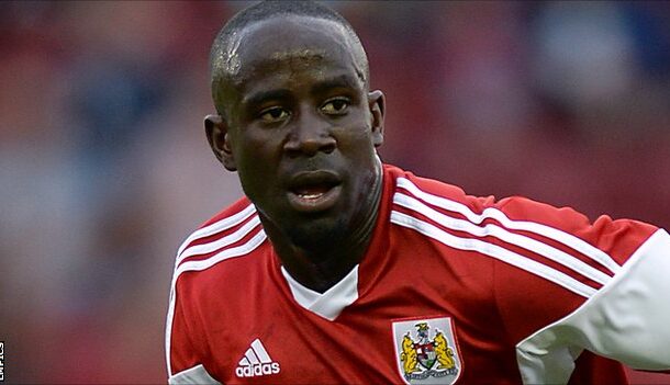 Action shot of a footballer in a red jersey with white stripes, running on the field and looking focused.