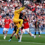 Soccer goalkeeper in a yellow kit leaps to punch or catch the ball while red-clad players challenge nearby on a green field, with a packed stadium in the background.