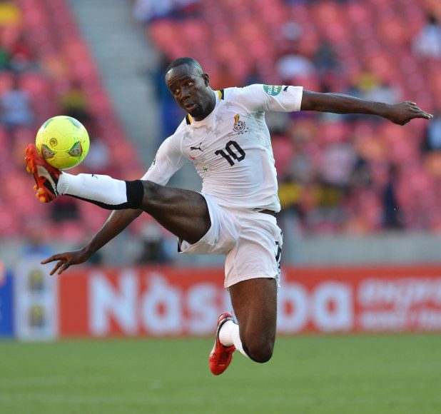 Ghanaian footballer wearing number 10, in white kit, jumping to kick a bright yellow ball in a stadium match