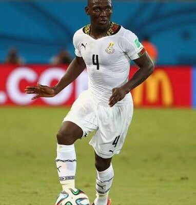 Ghana national footballer wearing white kit with number 4 dribbling a colorful soccer ball on a green pitch during a match, stadium blurred in the background.