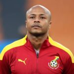 Head-and-shoulders portrait of a bald man wearing a red and yellow Ghana Football Association jacket, with the badge on the left chest, looking straight ahead.
