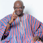 Smiling man with glasses, pointing his finger toward the camera while wearing a red-and-blue striped garment.