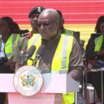 President delivering a speech at a podium with microphones, wearing a yellow safety vest and sunglasses, with officials behind him and a presidential seal on the lectern.