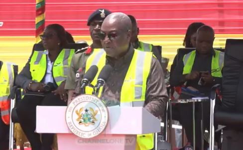 President delivering a speech at a podium with microphones, wearing a yellow safety vest and sunglasses, with officials behind him and a presidential seal on the lectern.