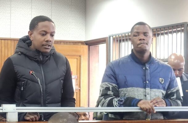 Two men stand behind a railing in a courtroom; one in a black puffer vest at a microphone, the other in a blue-gray jacket, with a police officer in the background.
