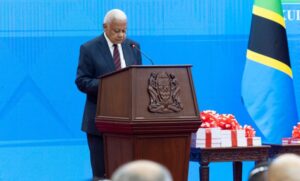 Older man in a dark suit delivers a speech at a wooden podium with a crest, against a blue backdrop and a Jamaican flag nearby, with gift boxes on a table beside him.