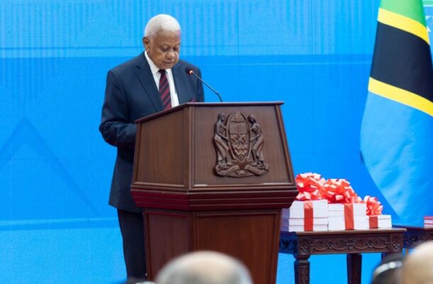 Probe | Ghanaguardian.com Older man in a dark suit delivers a speech at a wooden podium with a crest, against a blue backdrop and a Jamaican flag nearby, with gift boxes on a table beside him.