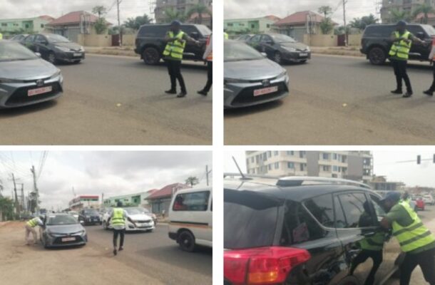 Police officer in a neon safety vest directing traffic beside a gray car on a city street; other vehicles are nearby.