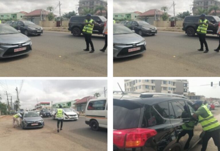 Police officer in a neon safety vest directing traffic beside a gray car on a city street; other vehicles are nearby.