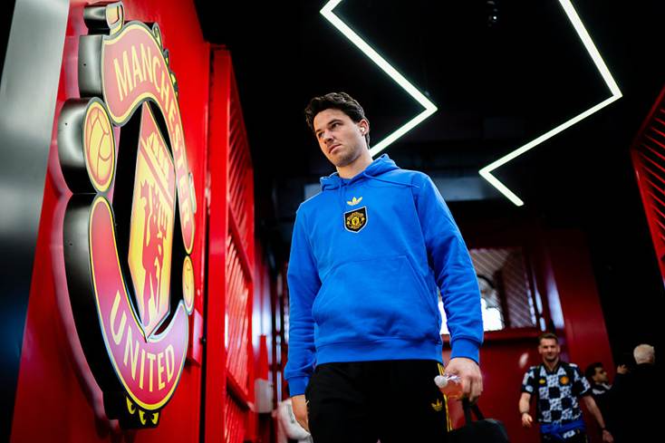 Man wearing a blue Manchester United hoodie walks through a hallway beside a large club crest on the left wall, neon shapes overhead.