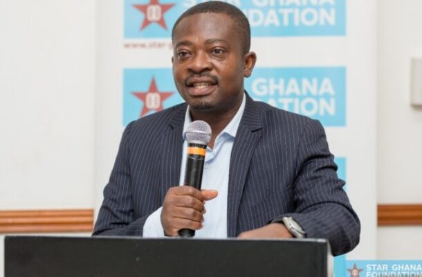 Man in a pinstriped suit speaks into a microphone at a podium, with blue Ghana Foundation banners in the background.
