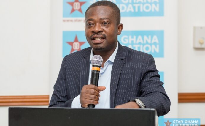 Man in a pinstriped suit speaks into a microphone at a podium, with blue Ghana Foundation banners in the background.