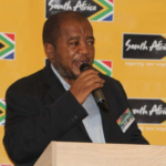 Man in a dark suit speaking into a microphone at a podium, with a bright yellow backdrop featuring South Africa logos.