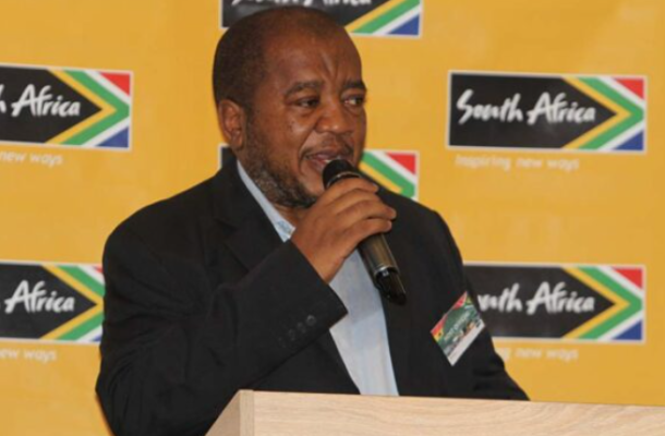 Man in a dark suit speaking into a microphone at a podium, with a bright yellow backdrop featuring South Africa logos.