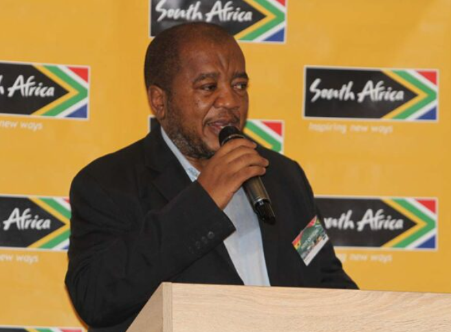 Man in a dark suit speaking into a microphone at a podium, with a bright yellow backdrop featuring South Africa logos.