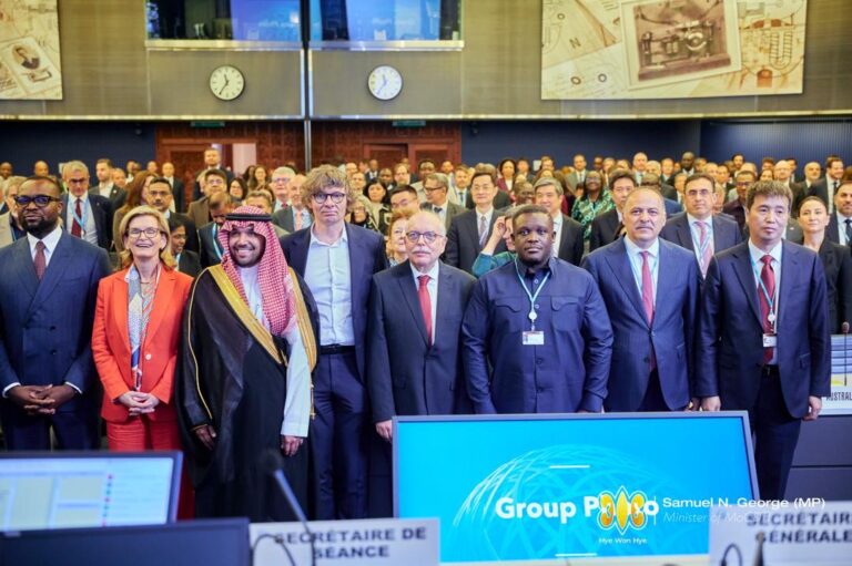 Group of formally dressed delegates posing for a photo at an international conference, with a blue display screen in front.