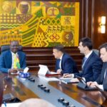 Officials in suits sit around a long conference table in a wood-paneled room; a man on the left holds a small flag while others work on tablets and documents, with a colorful wall art and a Ministry of Finance seal in view.