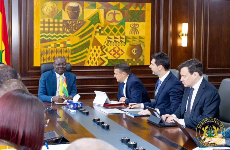 Officials in suits sit around a long conference table in a wood-paneled room; a man on the left holds a small flag while others work on tablets and documents, with a colorful wall art and a Ministry of Finance seal in view.
