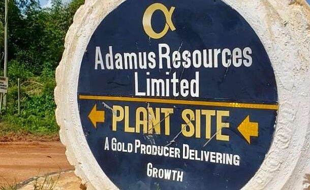 Round rock sign reading 'Adamus Resources Limited' with 'Plant Site' and 'A Gold Producer Delivering Growth' and arrows pointing left and right.