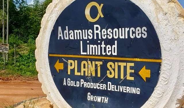 Round rock sign reading 'Adamus Resources Limited' with 'Plant Site' and 'A Gold Producer Delivering Growth' and arrows pointing left and right.