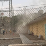 Emergency responders near a beige building inside a fenced electrical substation, as dust or smoke rises from the area.