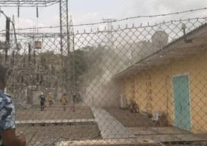 Emergency responders near a beige building inside a fenced electrical substation, as dust or smoke rises from the area.