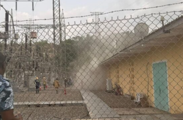 Emergency responders near a beige building inside a fenced electrical substation, as dust or smoke rises from the area.