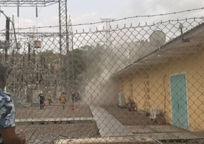 Emergency responders near a beige building inside a fenced electrical substation, as dust or smoke rises from the area.